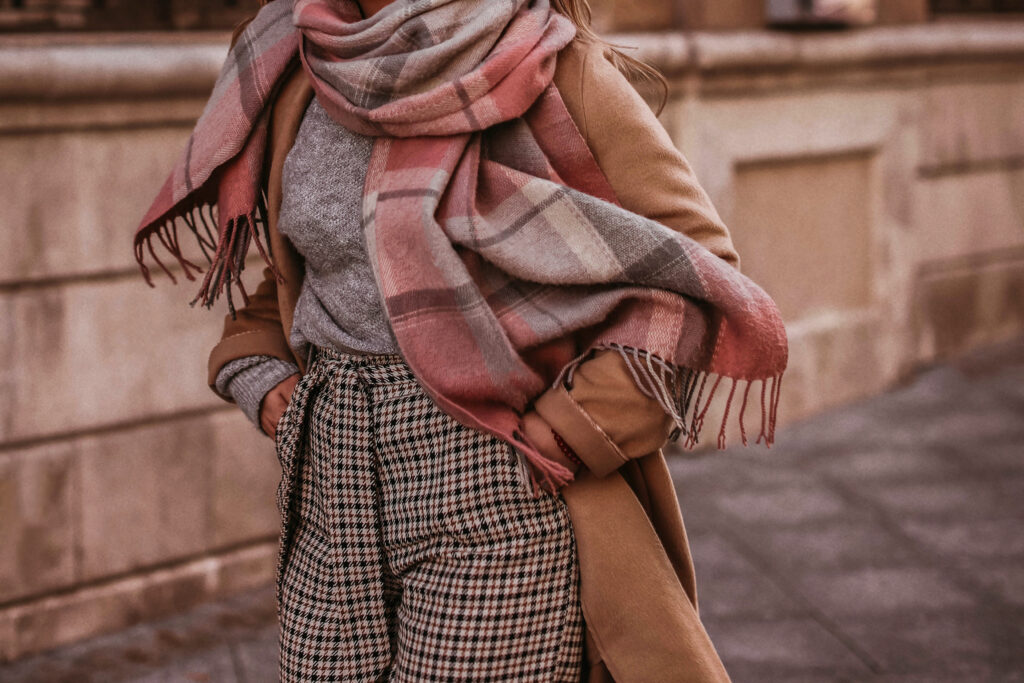 Woman in autumn stylish fashion brown long coat, scarf and plaid pants walking in the city. Female casual street style outfit. Trendy w dzianinach